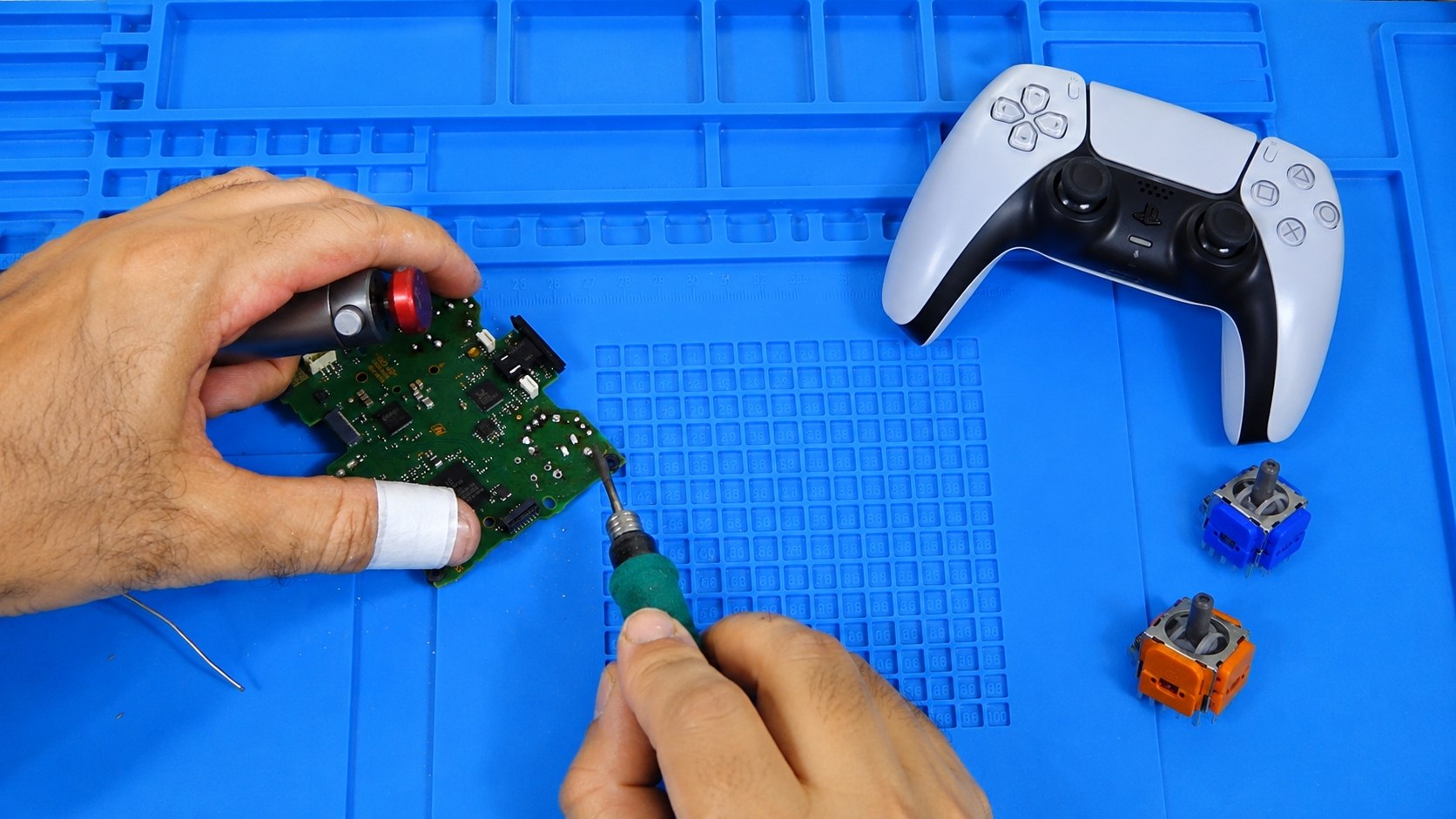Manos reparando una placa de un mando DualSense con soldador de aire caliente, mando PS5 y sticks Hall-effect al lado